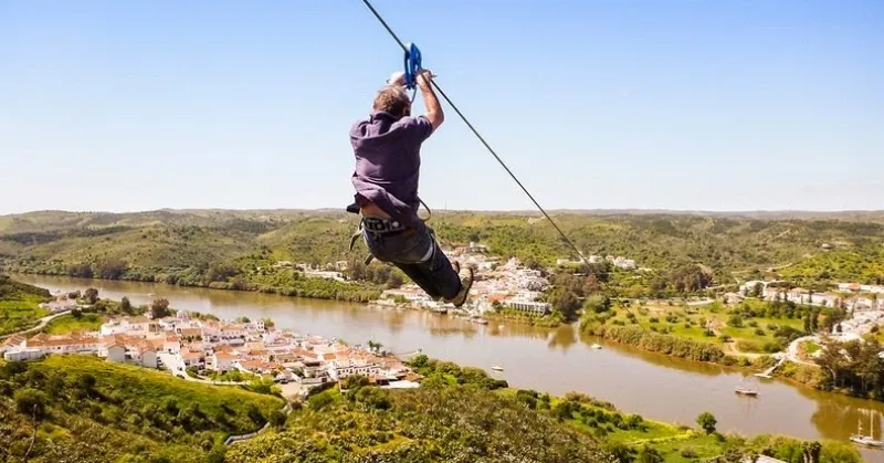 An adventurer sliding across the Guadiana River on the Limite Zero cross-border zipline connecting Sanlucar de Guadiana in Spain to Alcoutim in Portugal.